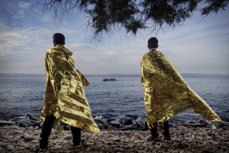 Dois homens recém-chegados à Ilha de Lesbos, na Grécia, usam cobertores sintéticos para se aquecer, tendo, ao fundo, barco com refugiados sírios que partiu da costa da Turquia © Espen Rasmussen