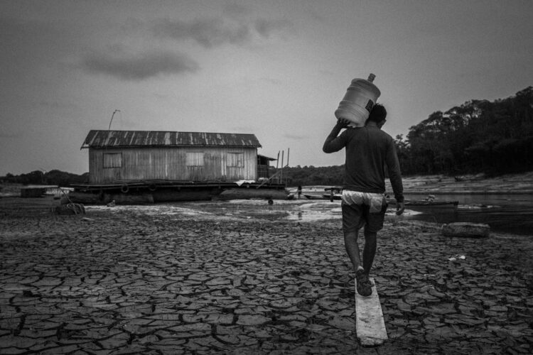 Jonathan Siqueira, 33 anos, carrega uma garrafa de água até o flutuante em que vive e que que encalhou no Rio Tarumã-Açu, um dos principais afluentes do Rio Negro, em Manaus, Amazonas, Brasil. Jonathan, que mora com outras nove pessoas na casa, disse que a falta de água foi o problema mais difícil da seca de 2023. O rio Negro, maior afluente do rio Amazonas, atingiu a cota de 12,70m em 2023, marcando a estiagem mais forte da história de Manaus e de todo o Amazonas:  comunidades ficaram isoladas, a navegação foi prejudicada, a distribuição de produtos caiu drasticamente e mais de 600 mil pessoas conviveram com dificuldade no acesso a água.