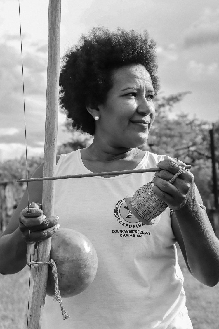 A retratada* posa segurando os principais instrumentos da capoeira: berimbau, caxixi e baqueta. O registro foi feito no pátio aberto do CEFOL – Centro de Folclore de Caxias – MA, na data de 11 de junho de 2022. A encenação foi proposital, parte para a realização do conjunto fotográfico.
*Rosemary Almeida, mulher, negra, brasileira, capoeirista
