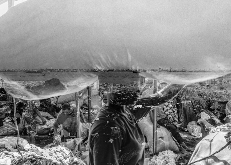 A portrait of a kayayei carrying a heavy sack of second-hand clothes, overlaid with the polluted shores of Jamestown Beach in Accra—a powerful reflection of how Western consumption continues to exploit both people and the environment in the Global South. Some kayayei carry bales as heavy as 55 kg, leading to severe injuries to their spines and necks.