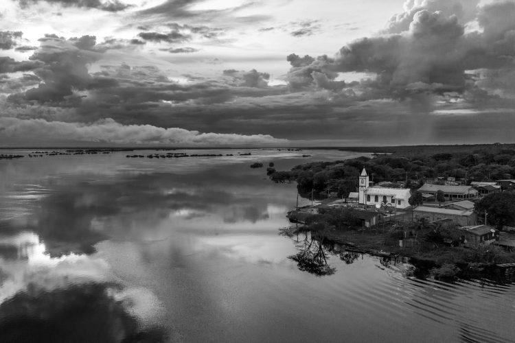 Essa imagem, feita com drone, nos mostra a comunidade de Carvoeiro onde a igreja domina a cena dos homens, disputando o protagonismo com a imensidão do rio e das nuvens.