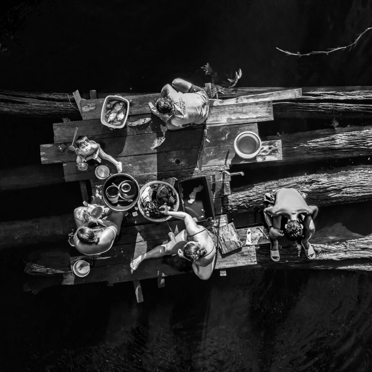 Nessa imagem o drone mostra varias atividades que acontecem nessa balsa nesse momento. Uma pessoa lava a roupa, outra a louça, outra limpa o peixe, outro escovas os dentes enquanto a criança flagra o drone. Conheça os finalistas do Prêmio Portfólio FotoDoc 2025