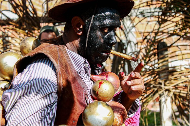 O Caçador - Com olhos atentos e o corpo coberto por símbolos de guerra, o Caçador é aquele que conhece a mata, o cheiro da fuga e o silêncio da resistência. Entre o fumo e os frutos, carrega a memória de quem persegue... e de quem já foi perseguido.