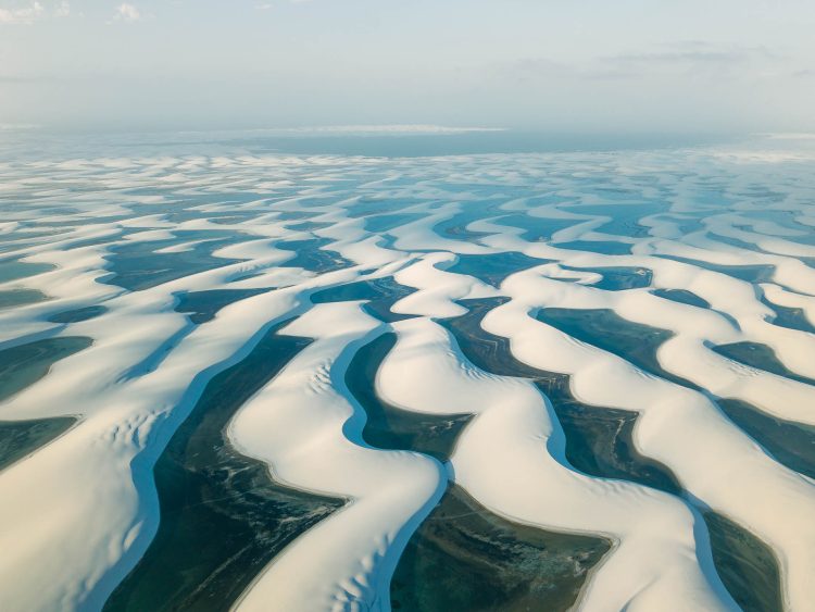 Arquipélago
Drone oblíquo, fim de tarde. 155 mil hectares de dunas brancas intercaladas por lagoas temporárias. Padrão se repete até horizonte: cada lagoa tem ciclo próprio — algumas já secam, outras atingem máxima profundidade. Não há testemunha humana visível. Apenas
geografia que se transforma em ritmo próprio. O grandioso não precisa de escala. Existe independente. Mas fotos seguintes introduzirão o mínimo corpos que atravessam este infinito, medindo com passos o que números não capturam.
Lençóis Maranhenses.
