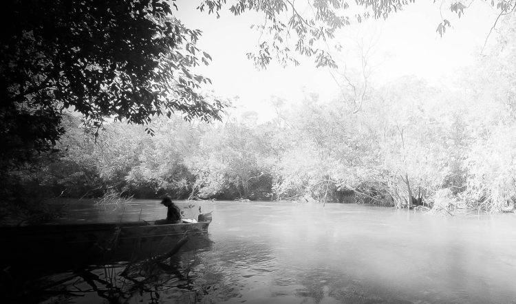 Caronte
Um dos homens aguarda pela chegada do outro em seu bote. Prepara-se para partir rio adentro, rumo a algo. Aqui, as sombras que o cercam e encobrem só não são mais opressivas que o branco do rio e da vegetação que o aguardam.